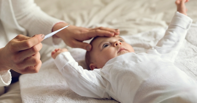 Bebé acostado sobre una cama, una mano sobre su cabeza y en la otra mano, un termometro